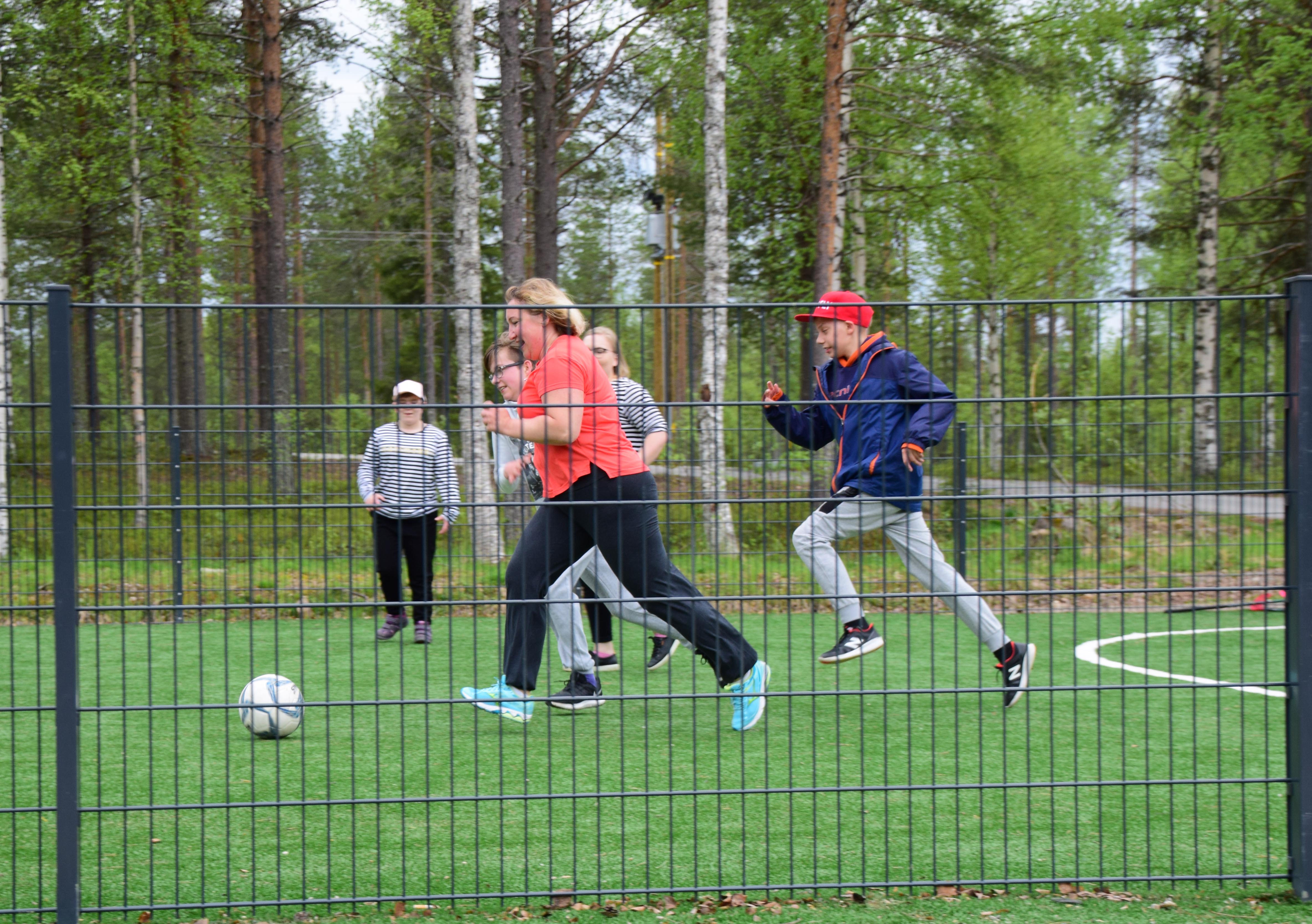 Nuoret pelaamassa jalkapalloa aidatulla tekonurmikentällä, taustalla metsää.