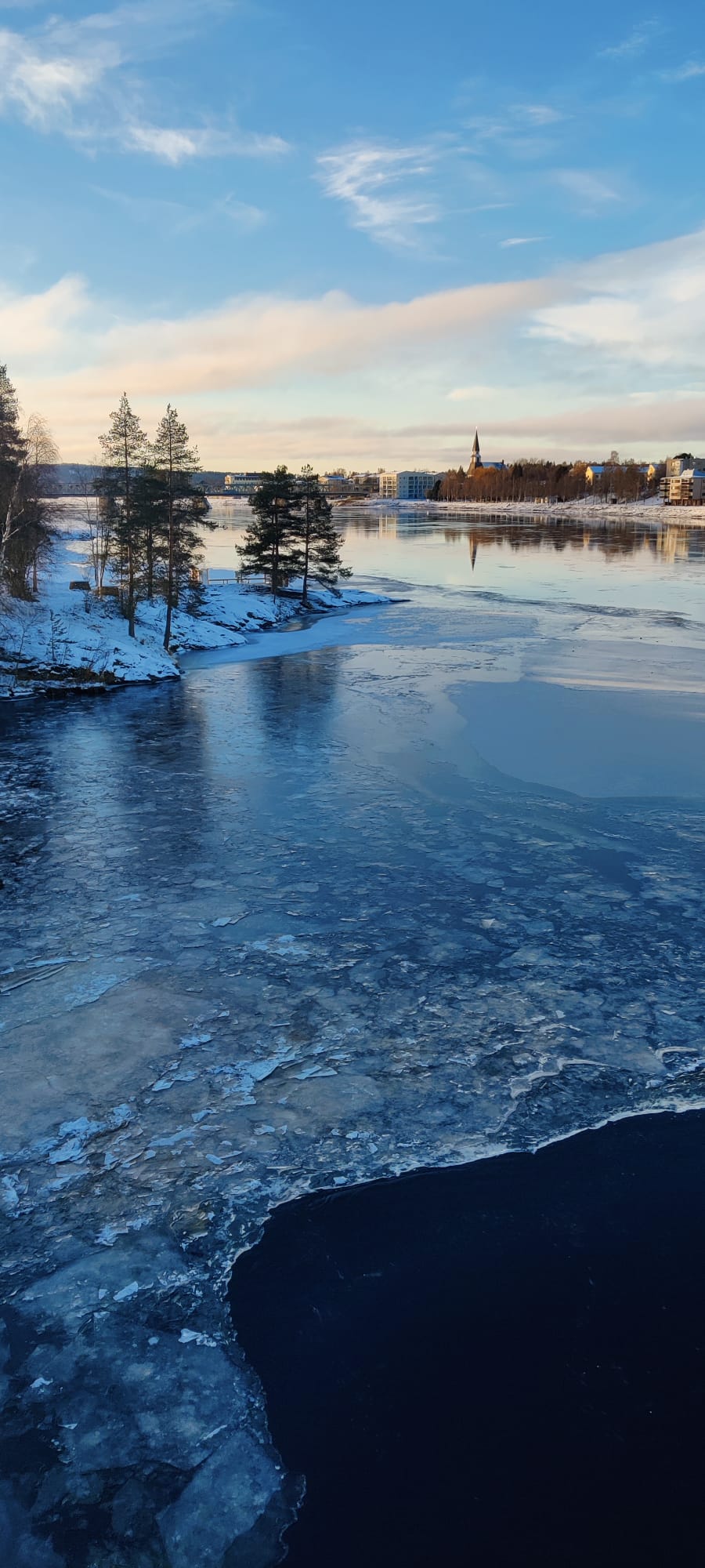 Kuvassa näkyy joenranta ja joen pinnalla jäätä monessa eri muodossa etutalalla sekä vaaramaisemaa ja kirkko...