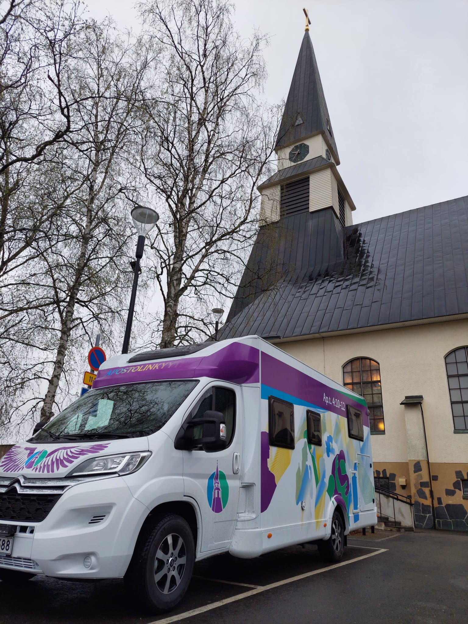 Kuva Apostolinkyyti-matkailuautosta ja sen värikkäistä teippauksista Rovaniemen kirkon vieressä.