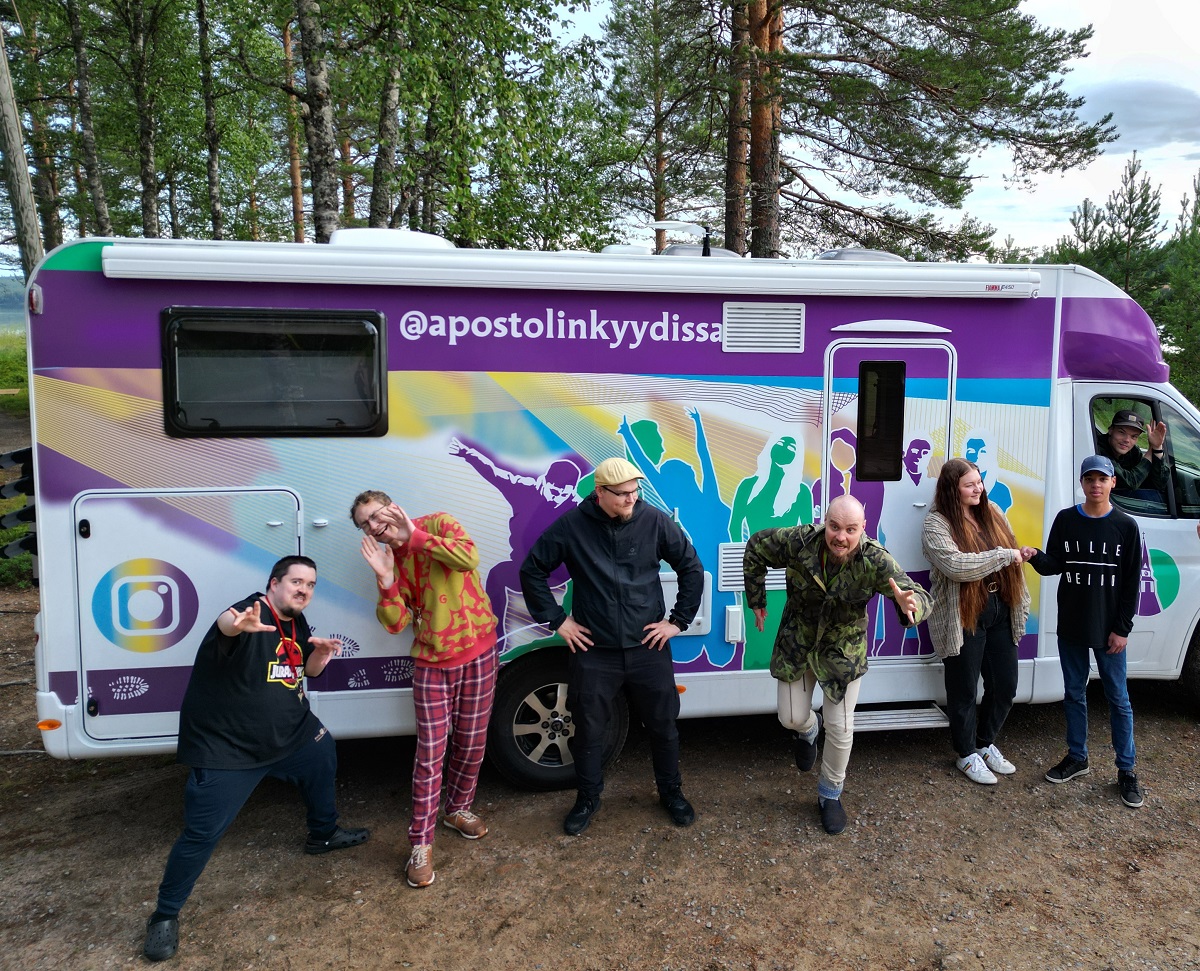 Nuorisotyönohjaajat ja yksi nuori poseeraavat liikkuva nuorisotila Apostolinkyydin edessä. Taustalla mäntyjä.